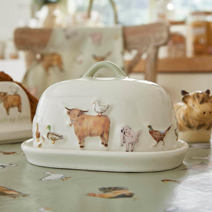 A white ceramic butter dish with a matching lid, decorated with painted farm animals including a cow, goose, duck, chicken, and rabbit. The dish is shown on a patterned tablecloth in a kitchen setting, with other matching animal-themed dishes blurred in the background.