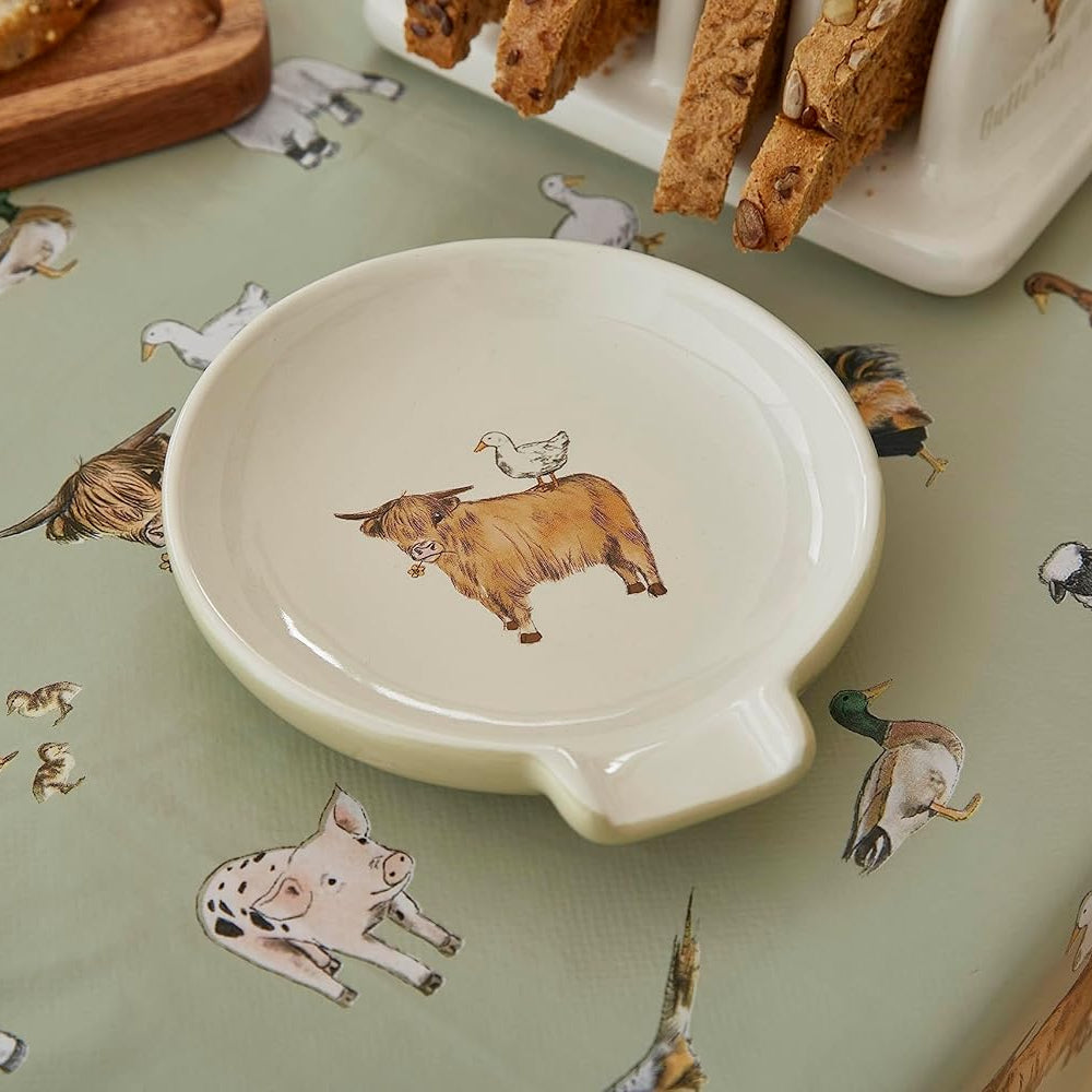 A small round ceramic dish with a handle, decorated with a brown cow illustration in the center, set on a sage green tablecloth printed with various farm animals.