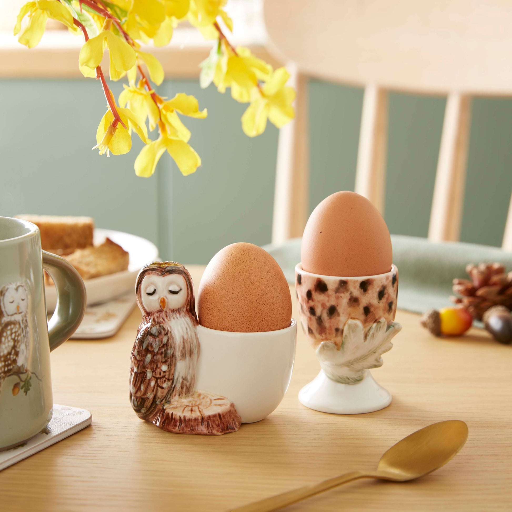 Cartco set of two ceramic egg cups featuring a sculpted owl and an acorn design on a wooden breakfast table