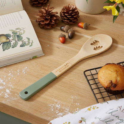 Cartco Cooksmart Forest Fable bamboo spoon with sage green handle and woodland leaf cutouts on a wooden kitchen table