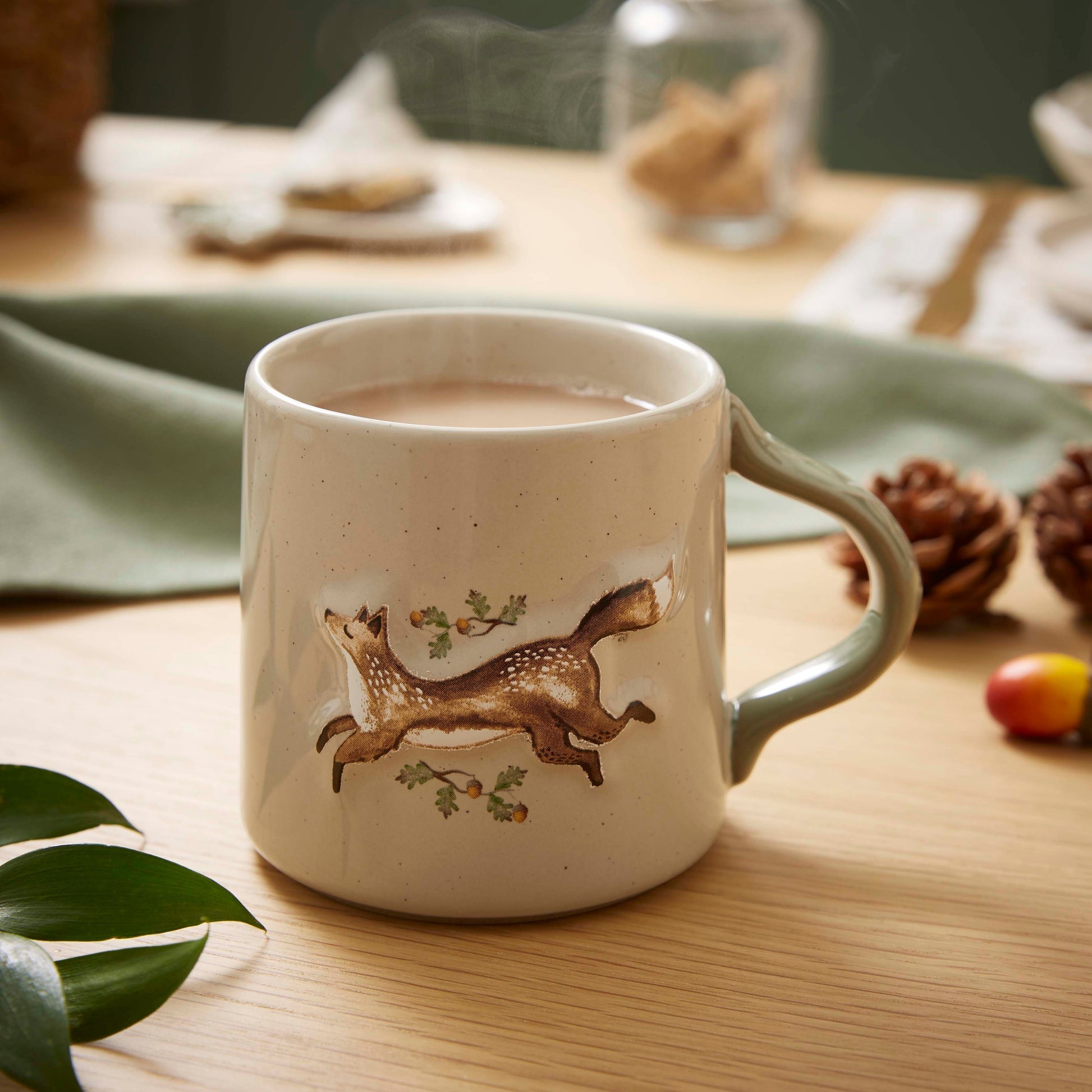 Cartco Cooksmart ceramic mug with a running fox, oak leaves, and acorns on a speckled cream body with a sage handle.