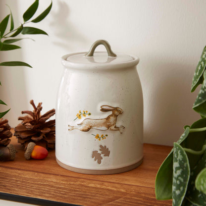 Cartco ceramic canister with a running hare, golden flowers, and a sage green loop handle on a wooden shelf.