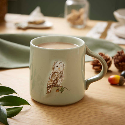 Cartco sage green ceramic mug featuring a peaceful owl illustration on an oak branch, set on a wooden table.