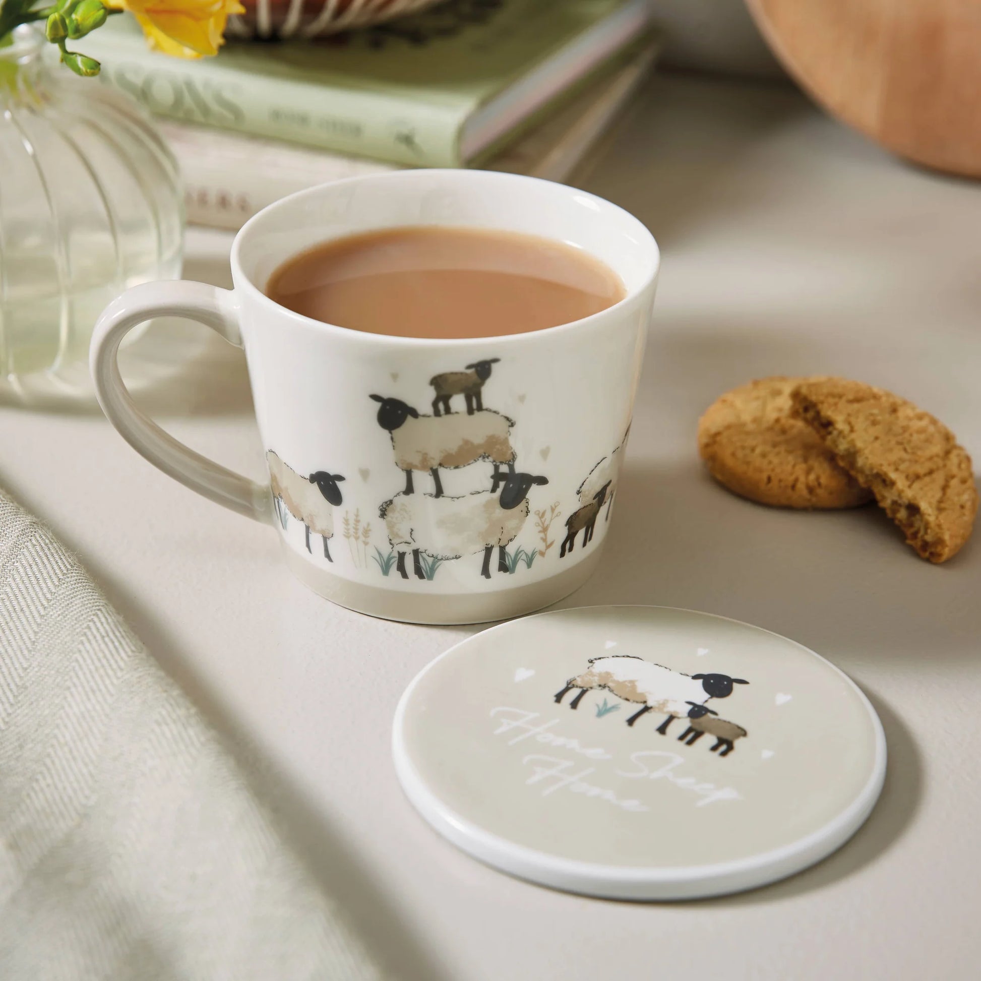 A white ceramic mug filled with tea, decorated with a sheep illustration, sits on a tabletop beside its matching lid. A cookie, books, and a glass vase are softly blurred in the background.